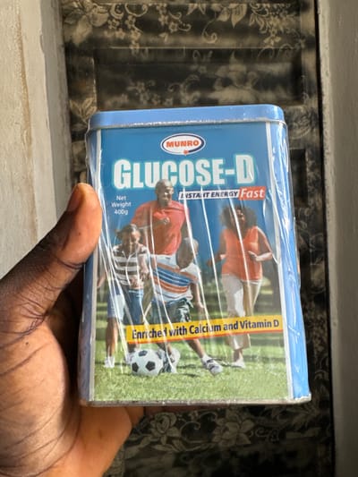 A person's thumb and index finger are holding a blue tin of "Glucose-D" drink powder. The tin is wrapped in clear plastic. The tin has an image of a family playing soccer on a grassy field. The text on the tin indicates "Instant Energy Fast" and "Enriched with Calcium and Vitamin D". The background of the image is a wall with a patterned wallpaper. The scene is indoors. It is not possible to determine the specific location within Awka, Nigeria from the image.
