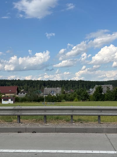 This is a landscape image, rotated 90 degrees clockwise. The scene depicts a roadside area with a guardrail on the right side, separating the road from a grassy verge and a dense forest. Beyond the forest, several houses with red and grey roofs are visible, suggesting a rural or suburban setting. The sky above is a bright blue, dotted with large, fluffy white clouds, indicating a clear, sunny day. The overall impression is one of a serene, rural landscape under a bright sky. The image doesn't contain any visible text or specific activities taking place. The time of day appears to be daytime, likely afternoon given the bright sunlight. The location context provided is Лугаваслабадскі сельскі Савет, Belarus.