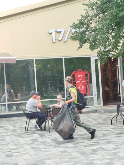 A man in camouflage pants and a yellow vest walks past a cafe in Zavodoukovsk, Russia, carrying a large black trash bag. He is walking on a paved outdoor seating area in front of a building with large windows displaying merchandise on shelves. Two men and a boy are seated at a table inside the cafe, reflected in the window. Another man is standing behind them. The building has "17/29" displayed in large white letters on its facade. A red sign with text in Cyrillic is visible in the window. The ground is paved with grey and beige square tiles.