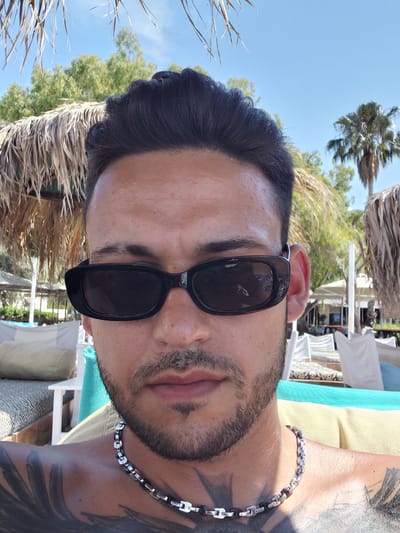 A close-up selfie of a man wearing black sunglasses and a silver and black chain necklace. He has dark hair styled upwards and stubble on his face. He is shirtless, revealing tattoos on his shoulder and chest. The background is a sunny outdoor setting with palm trees, thatched umbrellas, lounge chairs, and what appears to be a beach resort. The man is outdoors, likely on vacation in a warm, tropical or Mediterranean climate, given the vegetation and beach resort setting. The location is specified as the Municipal Unit of Diakopto, Greece.