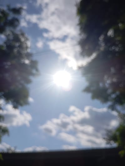The image, taken in Sozonivka, Ukraine, is a blurry, overexposed photograph of the sky on a sunny day. The sun is the brightest element, positioned centrally and radiating light in a starburst pattern. Fluffy white clouds are scattered across a bright blue sky. Dark, out-of-focus foliage frames the top edges of the image, suggesting trees on either side. The bottom of the frame is dominated by a dark, blurry horizontal band, possibly representing a roofline or a dark surface. No people or specific structures are discernible due to the lack of focus and overexposure.