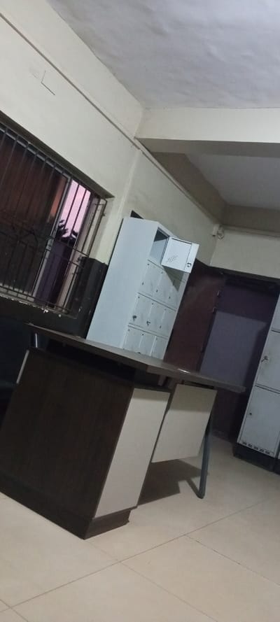 An interior shot features a tilted perspective of an office space. A dark wood and white desk anchors the scene, with a tall, light gray locker unit behind it, one door ajar. Adjacent to the locker is a doorway. Bars cover the nearby window, revealing a glimpse of the exterior sky. The floor consists of square tiles. The room is lit by indoor artificial light, casting shadows that emphasize the angled view.