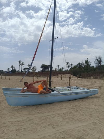The image shows a shirtless man lying on a light blue sailboat pulled onto a sandy beach. He is wearing bright orange swim trunks and flip-flops. The man is resting his head on his hand, with his legs bent. His pose appears relaxed and somewhat playful.
The sailboat is a catamaran-style dinghy with two hulls. It has a tall mast with a sail that is partially furled and features red, white, and blue stripes. The boat is positioned on the sand, facing towards the viewer and slightly to the left.
In the background, a line of palm trees and other vegetation suggests a coastal or tropical environment. A fence made of natural materials runs along the edge of the beach, separating it from the vegetation. The sky is partly cloudy with fluffy white clouds against a bright blue background, indicating it is daytime and the weather is fair. The sand appears dry and lightly textured with some footprints visible. The scene evokes a sense of leisure and a beach vacation.
The location is identified as Sagunto, Spain. There is no visible text in the image.