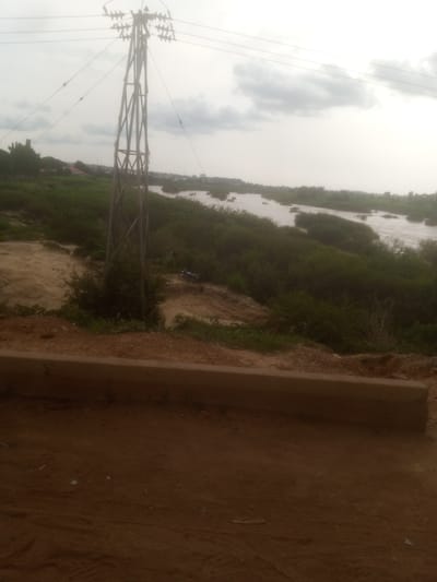 This is a full-frame, eye-level, outdoor, medium shot of a wide, shallow river with several small islands covered in lush green vegetation. The river appears to be in a flood state, with water flowing rapidly around the islands. In the foreground, there is a dirt path with a low, brown concrete barrier running horizontally across the frame. Behind the barrier, more dirt and scattered green vegetation are visible. To the left of the frame, a tall, metal electricity pylon stands prominently, with multiple wires extending outwards and upwards into the overcast sky. In the middle ground, behind the pylon and along the riverbank, dense green foliage lines the water. A dark-colored motorcycle is parked on the dirt path behind the pylon. The background shows a hazy, distant treeline and structures suggesting a settlement. The overall environment suggests a rural or semi-urban area near a body of water in Nassarawa, Nigeria, under a cloudy sky.