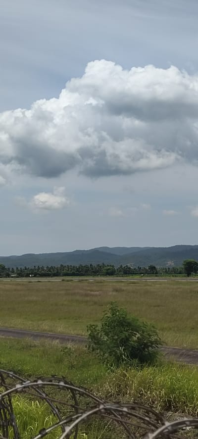 A vertical, medium-range shot captures a vast expanse of dry grass and brush in the foreground, transitioning into a paved path or road. Beyond this lies a flat, open field, hinting at an airfield or large clearing, with a distant line of lush, green trees and rolling hills forming the horizon. The sky above is filled with dramatic, volumetric clouds, ranging from bright white to deep gray, suggesting an overcast or approaching storm. The scene appears to be an outdoor environment, possibly a rural or undeveloped area within Buengbakjok, Indonesia. The foreground features a barbed wire fence, partially obscuring the view of the field. No people or animals are visible.