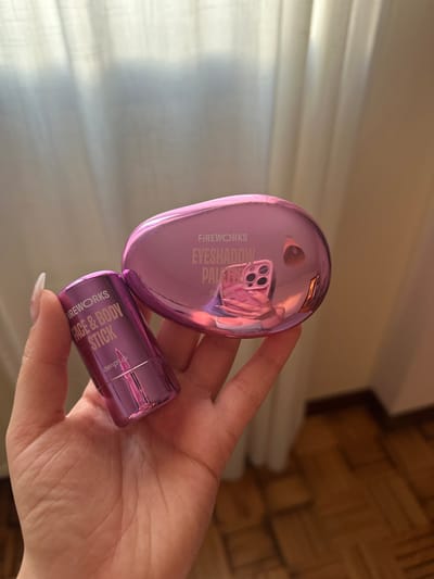 A close-up, eye-level shot depicts a person's fair-skinned left hand holding two metallic magenta-colored cosmetic products. The hand, exhibiting a light-colored manicure on the visible fingernails (thumb and index finger), securely grips both items.

On the left, a cylindrical "FIREWORKS FACE & BODY STICK" by "deliplus" is held vertically, displaying an arrow pointing downwards. To its right, an oval-shaped "FIREWORKS EYESHADOW PALETTE" by "deliplus" is held horizontally. The highly reflective surface of the eyeshadow palette prominently features an inverted reflection of a smartphone, identifiable by its multi-lens camera array, encased in a pink cover. The hand holding the smartphone and a portion of the photographer's upper body, wearing a purple garment, are distinctly visible in this reflection.

The background is divided into two main sections. The upper portion consists of light-colored, possibly off-white or cream, vertically pleated curtains, suggesting the presence of a window and contributing to the soft, diffused lighting. The lower right portion of the background reveals a wooden parquet floor, laid in a patterned arrangement, along with a dark brown or black baseboard where the wall meets the floor.

The visual information within the image does not provide any specific clues or landmarks to determine the exact location within the city of Gijón/Xixón, Spain.