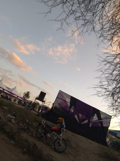 The image shows a motorcycle parked in front of a large black and white tent structure with purple fabric draped over it. The tent is located in an open area with sparse vegetation and a dirt ground. The sky is clear with faint clouds. The motorcycle is a red and black model with a yellow helmet on the seat. In the background, there are other tents, some of which are white, along with people and a sound system on a platform. This scene is located in Athi River, Kenya, at coordinates latitude -1.457 and longitude 36.975. The image appears to have been taken at dusk.