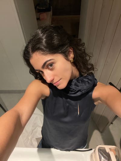 A young woman with dark curly hair and fair skin poses in a selfie. She wears a navy blue silk top with a small slit at the neckline. Her right arm is raised, drawing attention to her smooth skin. She has green eyes and a gold earring in her left ear. The background includes a white countertop, a makeup bag, and wooden paneling. The photo is slightly rotated, giving it a casual and off-center feel.