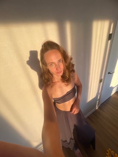 A young woman with medium-length, wavy brown hair and blue eyes is taking a selfie in a room with a textured wall and wooden floor. She is wearing a dark grey bandeau top and matching wide-leg pants. The room is illuminated by natural sunlight casting shadows from a window onto the wall. A white door with a black handle is visible on the right side of the image. There is a floral arrangement partially visible in the bottom right corner. The scene appears to be a domestic interior in the city of Podshivalovo, Russia.