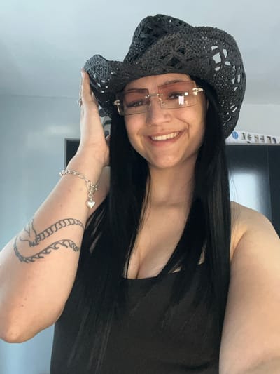 A woman with long, dark hair wears a black, crocheted hat and rimless sunglasses. She is smiling and looking towards the camera. A silver bracelet with a heart-shaped charm adorns her wrist, and she has a tattoo on her arm. The scene appears to be indoors, with a plain wall in the background. The lighting suggests it is daytime.