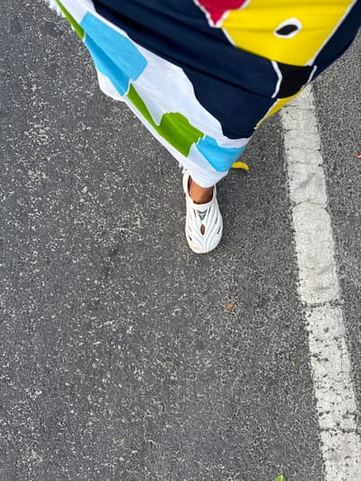 A downward-facing shot taken on an asphalt surface, possibly a street, depicts a person's leg and foot adorned with a white, sandal-like shoe with cut-out designs. The person is wearing a brightly colored garment featuring shades of blue, green, white, yellow, red, and black. A white line is visible on the asphalt, situated near the lower left corner of the frame. The location appears to be Jimbaran, Indonesia.