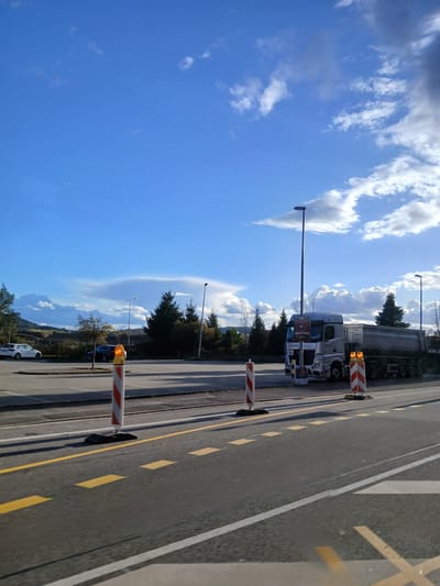 The image depicts an outdoor scene in Homburg, Germany, under a partly cloudy blue sky with bright sunlight.

In the immediate foreground, a paved asphalt road surface is visible, marked with yellow dashed lines and a solid white line on the right, along with several yellow 'X' markings towards the bottom right corner. Two red and white striped construction barrier posts, each topped with an orange flashing light, are present: one prominently in the left foreground and another further right, closer to the midground. Both posts rest on dark, possibly rubber, bases.

The midground features a paved area resembling a parking lot or wide shoulder on the left, where a white sedan or SUV and a dark-colored sedan or SUV are parked. Further right, a light grey Mercedes-Benz Actros tractor unit is parked, coupled with a matching grey tandem-axle dump or bulk goods trailer. The truck's cab displays "NEUMANN" branding and several attached signs, including one that appears to relate to "ZBF Homburg" and another indicating "NEUMANN Entsorgung & Services GmbH." A third red and white striped barrier post with an orange flashing light stands in front of the truck. Behind the parking area and truck, a line of evergreen trees (conifers) is visible, interspersed with deciduous trees. Several tall black streetlights are positioned throughout this area.

The background on the left reveals rolling hills or elevated terrain covered in sparse vegetation, suggesting a more natural landscape beyond the constructed area. The sky above is predominantly bright blue with scattered white cumulus clouds, some forming larger, darker masses towards the horizon. No individuals are visible in the scene. The vehicles are stationary.