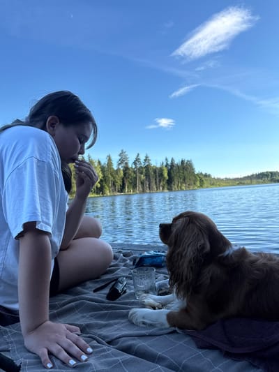 A young person with dark hair and a white t-shirt sits on a dock, facing a brown and white dog. The person is holding a small object to their mouth. The dog is lying down, looking at the person. The dock is covered with a gray blanket. There is a glass and a black object on the blanket. The background shows a lake with trees and a blue sky with clouds. The location is the city of Zamosh'e (Zaklinskoe S/P), Russia.