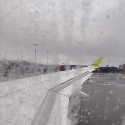 The image depicts a view from the window of an aircraft, looking outwards onto an airport tarmac on an overcast day. The windowpane is covered with numerous visible raindrops, causing a diffuse, blurred effect, particularly in the immediate foreground.

In the mid-ground, a significant portion of a white aircraft wing is visible, extending diagonally from the lower left towards the right. The wing features a distinct lime green wingtip, on which the black text "airBaltic" is clearly legible. Partial text reading "NO STEP OUTSIDE THIS" is discernible on the flat surface of the wing.

Beyond the aircraft wing and the wet tarmac, airport infrastructure is present under a grey, cloudy sky. A dark-colored terminal building or structure in the background prominently displays the capitalized word "RĪGA". To the left of "RĪGA", another structure, possibly a jet bridge or part of the terminal, shows the branding "Revolut". Several tall, grey light poles are scattered across the airport environment. The tarmac appears wet, consistent with the rainy conditions indicated by the window. The scene is located at an airport in the city of Skulte, Latvia. No individuals are explicitly visible. The aircraft appears stationary.