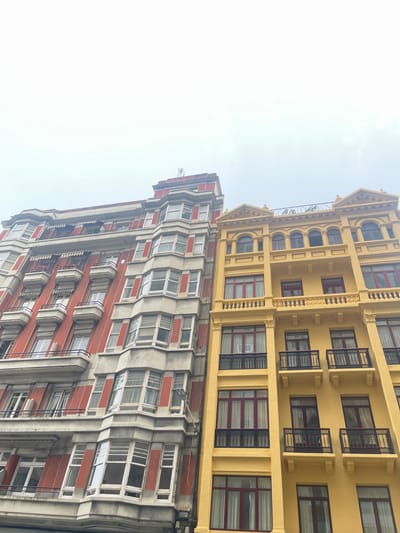 This media file shows a low-angle view of two adjacent buildings in A Coruña, Spain. The building on the right is a lighter yellow and features ornate architectural details, including decorative cornices, arched windows with dark frames and red curtains, and wrought-iron balconies. The building on the left is primarily reddish-orange with concrete accents, characterized by a more modern, stepped facade with numerous windows. These windows have white frames and some appear to have blinds. Both buildings are multi-story structures. The sky is overcast and bright, suggesting a cloudy day. There is no visible activity or people in the image, and no text is present. The scene is a street view of urban architecture.