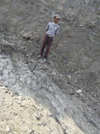 An adult male stands on a steep, excavated earthen slope in Hoche, Nigeria. The man, with dark skin, wears a light grey short-sleeved collared shirt with two dark shoulder stripes and a badge on the left breast pocket, dark brown trousers with a prominent red stripe down each outer seam, a light-colored belt, dark shoes, and a grey cap with a red band and a white emblem. His hands are clasped behind his back, and he gazes slightly to his left with a neutral expression. The surrounding environment consists of a rugged, light greyish-brown, loose soil and rock embankment displaying irregular textures, striations, and evidence of erosion or excavation. The ground beneath him is similarly composed of dirt, clay, and scattered debris.