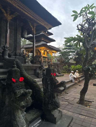 The image depicts a scene at a temple in Dawan, Indonesia. In the foreground, two dark stone dragon statues with red flowers on their heads are visible, one with its mouth open. To the right, two men in white traditional clothing are sitting on stone steps. Behind them, the temple architecture features multiple tiers of dark, thatched roofs with ornate golden detailing. The ground is paved with square stones, some of which are cracked or uneven. Trees and lush greenery surround the temple, suggesting an outdoor setting within a temple complex. The sky is overcast, and the lighting indicates it may be dusk or a cloudy day.