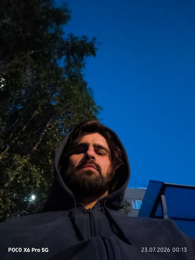 In the city of Kogalym, Russia, an individual with dark, medium-length hair and a beard is seen in a selfie, looking directly at the camera. They are wearing a dark hooded sweatshirt. The background consists of a tree on the left, a clear blue sky, and a building with blue awnings on the right. The timestamp "23.07.2026 00:13" and the text "POCO X6 Pro 5G" are visible in the lower corners of the image. The lighting suggests it is twilight or early night.