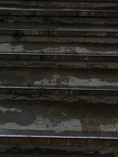 This is a close-up, overhead shot of a set of weathered concrete stairs, possibly outdoors in an urban or industrial setting. The stairs are dark gray and appear wet, with visible streaks of moisture and discoloration. The concrete surface is cracked in places, and there are patches of lighter gray material that might be remnants of paint or a different type of coating.

The lighting is dim, creating strong shadows between the vertical risers of the stairs, emphasizing their ribbed texture. The overall impression is one of decay and neglect. There are no people or distinct objects visible, only the stark, textured surface of the stairs. The scene feels somber and perhaps a bit oppressive due to the dark tones and the implied wetness, suggesting a recent rain or damp conditions. There is no visible text in the image. The context provided of Tikhoretsk, Russia, hints at a potentially cold or overcast environment.