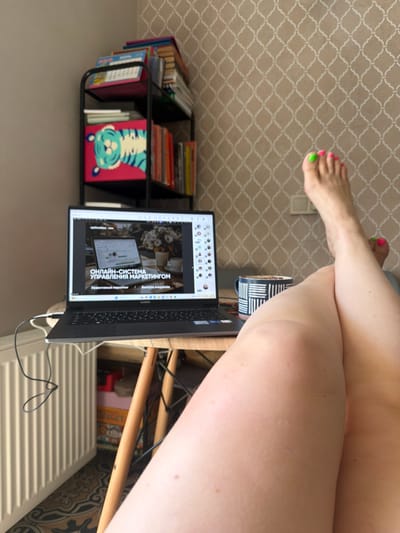 The image shows a person's legs and feet in the foreground, with their toes painted in bright pink and green nail polish. The person is sitting, with their legs crossed. In the background, there is a laptop open on a desk, displaying a presentation or website with Russian text that reads "ОНЛАЙН-СИСТЕМА УПРАВЛЕНИЯ МАРКЕТИНГОМ" (Online Marketing Management System) and "Эффективный маркетинг" (Effective Marketing). There are also several books and binders on a black shelf unit. The floor has a patterned wallpaper. The setting appears to be indoors, likely a home office or study. The lighting suggests it could be daytime. The overall mood is casual and productive.
