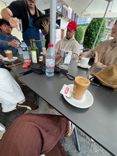The image captures a street-side cafe scene in Jerez De La Frontera, Spain. The shot is taken from an elevated angle, looking down at a dark gray table top. Several individuals are seated around the table, their bodies partially visible. A man wears a red baseball cap and a light-colored sweatshirt. Another person is wearing a tan jacket and a brown hat. Another person is wearing brown pants and brown and white shoes. A waiter, dressed in a black shirt and blue jeans, serves customers, holding a tray in his left hand and carrying a glass in his right hand. A second waiter wears a blue jacket and is holding a glass.
Various items are placed on the table, including glasses of coffee, water bottles, a bottle of olive oil, sugar packets, a wallet, and cellphones. The cafe is located outdoors, as suggested by the presence of an umbrella. A building facade and lush greenery can be observed in the background, indicative of a city street.