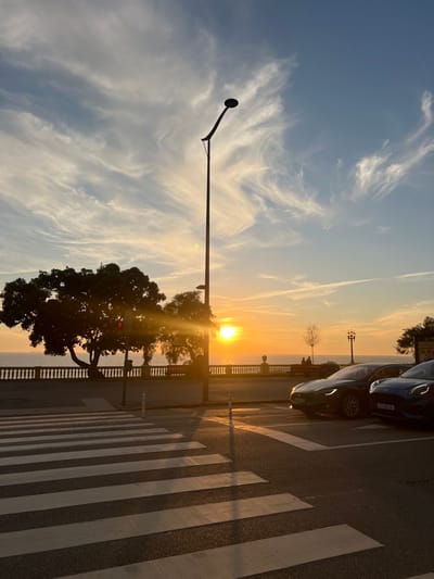 The image captures a coastal scene at sunset in Aldoar, Foz do Douro e Nevogilde, Portugal. The sun, a bright orange orb, is low on the horizon directly over the Atlantic Ocean, casting strong backlighting and long shadows. The sky above is a gradient from deep orange at the horizon to clear blue overhead, populated by thin, wispy white cirrus clouds.
In the foreground, a white-striped pedestrian crosswalk traverses a dark asphalt road. To the right of the crosswalk, two vehicles are parked facing right. The closer vehicle is a dark grey Tesla, with its front headlights illuminated. Adjacent to it, a blue Ford Puma displays the license plate "AT 58 UV" and also has its headlights on.
A tall, dark modern street light, featuring a curved arm and a circular light fixture, stands prominently in the midground, silhouetted against the sun. To the left, a large, dense tree is heavily silhouetted, partially obscuring the sun's brilliance. Along the coastline, a white stone balustrade with decorative pillars extends horizontally. Behind the balustrade, on a promenade, several indistinct human figures are visible in silhouette, with at least two individuals seated and others standing. Further right, another traditional-style street lamp is visible. The water surface appears calm and dark under the setting sun.