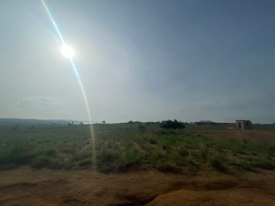 The image depicts a bright, sunny day in Buga da Gindi, Nigeria. The foreground is dominated by a reddish-brown, unpaved surface, possibly a dirt road or cleared land. Beyond this, a vast, open field of green grass stretches into the distance. Scattered throughout the field are sporadic patches of taller, wilder grass and a few scattered trees.

In the middle ground, on a slightly elevated area of the field, are two structures. To the right, a small, rectangular building with a dark, open doorway and a simple roof is visible. To the left of this building, further in the distance, is a larger, more complex structure, possibly under construction or an unfinished building, with a visible framework and a partially covered roof.

The background reveals a hazy, distant landscape with rolling hills and what appears to be a body of water, likely a river or lake. The sky is bright and clear, with the sun prominently positioned in the upper left quadrant, casting a strong lens flare that streaks diagonally across the image and creates a hazy, washed-out effect in that area. The overall impression is of a rural or semi-developed area under a clear sky.