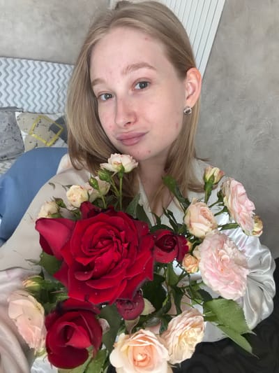 The image depicts a young adult female located in Rossiyskiy, Russia. She has shoulder-length blonde hair, light brown eyes, and fair skin with visible facial blemishes. She wears a small, silver-toned, irregularly shaped stud earring in her right ear. Her lips are lightly pursed, forming a subtle expression. She is dressed in a light-colored, possibly satin, garment with a faint floral pattern. She is holding a fresh bouquet of roses, featuring large deep red roses prominently, surrounded by smaller light pink and cream-colored roses and buds. Water droplets are visible on the petals and leaves. The background on the left includes a blue pillow, a grey and yellow patterned pillow, and a grey and white zigzag patterned fabric or headboard. The wall behind her on the right is light grey with a stucco texture. A white, vertically slatted structure, possibly a radiator cover, is visible in the upper right quadrant.
