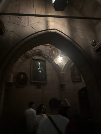 The image depicts an indoor scene with people queuing in a dimly lit, ornate hallway that appears to be part of an attraction or themed environment, potentially a museum or theme park. The architecture features stone walls with arched passages and classical-style paintings hanging on the walls. In the foreground, several individuals are partially visible, facing away from the camera and appearing to wait in line. The lighting is dramatic, casting shadows and highlighting architectural details. The specific location in Tongzhou District, China, is not discernible from the visual information provided.