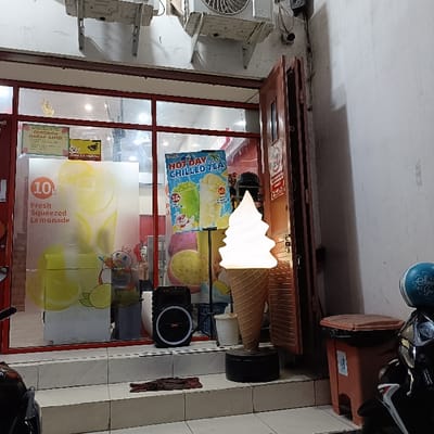 The scene is located in Kaliwungu, Indonesia, at coordinates -6.963 latitude and 110.251 longitude. The image shows a storefront with a large window display. The storefront is located on a sidewalk with a streetlight and a brown trash can. There is an advertisement on the window for "Hot Day Chilled Tea." The advertisement features images of iced tea in glasses with straws, and a cartoon character. The storefront is also adorned with a large, illuminated cone-shaped structure. The structure is white on the top and beige on the bottom and appears to be a cone, with a white, cone-shaped object sitting on top. There are several yellow lemons visible on the storefront, and a black speaker on the floor. The storefront also has a metal frame door leading inside. There are also what appear to be large, white ice cream cones, and a person in front of the storefront, but they are obscured by the white ice cream cone. The storefront is partially illuminated by a streetlight and there are a couple of people visible inside the storefront. The scene is lit by artificial lighting and appears to be taken on a night time or evening, as the lights are on in the storefront, and there is a street light. The scene is also characterized by a variety of colors, including bright yellow, red, white, brown, and green, with the image seemingly captured on a phone camera. There is a small area of space between the storefront and the trash can. There is a motorcycle parked in the space to the right of the storefront and the motorcycle is partially visible. The motorcycle is black and is partially obscured by the white cone-shaped object. The storefront is also adorned with small, decorative signs advertising "Fresh Squeezed Lemonade" and "10k." There is a green, plastic container visible in the background near the window, but it is partially obscured by the white cone-shaped object. There are no clear indications that anyone is interacting with anyone or anything in the image.