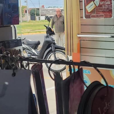 An older man with glasses and a striped shirt is standing next to a gray moped on a street in San Fernando del Valle de Catamarca, Argentina. The scene is viewed through a store window with various objects and advertisements displayed. A gift card and another advertisement are visible on the window. Beyond the street, there is grass, buildings, and a clear sky.