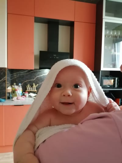 A baby, wrapped in a white towel, is being held by an unseen person. The baby is looking at the camera with a slight smile. The background appears to be a kitchen with orange cabinets. The location is in the city of Trubacheyevka, Russia.