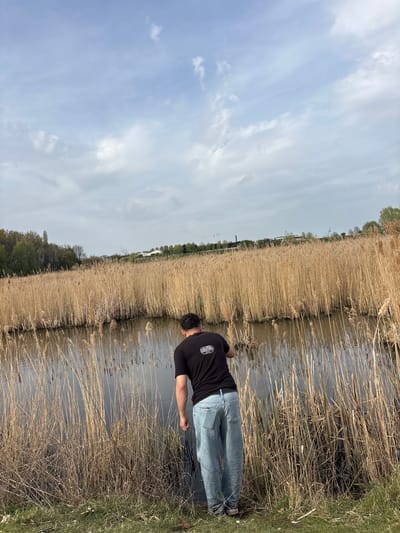 A man stands along a waterway. He wears a black shirt and light blue jeans. The waterway is bordered by tall reeds on the left and grass on the right. The sky is partly cloudy. The location is likely in Kruibeke, Belgium, based on the presence of similar landscapes.