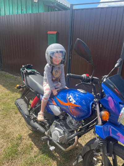 A young girl wearing a clear-visored helmet sits on a blue motorcycle parked on a grassy area in front of a brown metal fence. The girl is wearing a grey long-sleeved shirt with a cartoon image and patterned leggings. The motorcycle has orange accents and the "TVS" logo on the fuel tank. In the background, a green wooden structure with a small red and green mailbox is visible. The scene appears to be in a rural or suburban setting, possibly in the city of Сухринское, Russia, given the context. The sky is bright blue with some clouds. The ground is covered in dry grass and some patches of green vegetation.