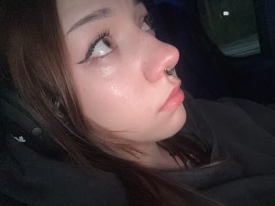 A close-up, profile view shows the face of a young person with brown hair. The person is wearing black clothing, and a silver septum ring is visible on their nose. The person's eyes are looking upwards, and there appear to be tears or a wet sheen on their cheeks. The background is dark, with a hint of a green and white reflective surface in the upper right portion, suggesting a window or skylight. The image is an indoor shot, possibly taken in a vehicle, given the reflective surface. The city of Medemciems, Latvia is not depicted in this image.