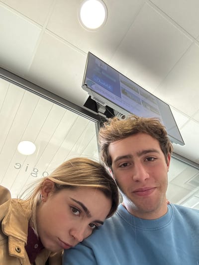 The image depicts a close-up selfie of two young adults inside the Imagerie Médicale du Grand Domaine, located in Saint-Priest, France. The female subject, on the left, has blonde hair and wears a tan jacket over a burgundy top; her head rests on the male subject's left shoulder, and she displays a slight pout. The male subject, on the right, has brown curly hair and wears a light blue crewneck sweatshirt; he looks directly at the camera with a neutral expression. Both subjects occupy the lower half of the frame, indicating a selfie perspective.

In the background, a white suspended ceiling with grid lines and a circular recessed light fixture is visible. A flat-screen television is mounted on the ceiling in the upper right, displaying the text "Doctolib" prominently, followed by "IMAGERIE MEDICALE DU GRAND DOMAINE" and "Saint-Priest," identifying the facility and its location. A clear glass partition extends across the mid-ground, behind the subjects, showing reflections, including inverted text that appears to be "eiga" or "eigam" which could be a reflection of "Imagerie". The overall environment indicates an indoor, clinical waiting area setting.