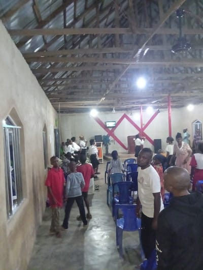 An indoor shot shows people gathering in what appears to be a church or meeting hall. The hall is minimally furnished with blue plastic chairs, and the walls are plain. Some individuals are standing or walking near the entrance area on the left, while others are congregated closer to a raised area at the back, where a television and red decorative elements are visible. The ceiling is exposed with wooden beams and a corrugated metal roof. The lighting is provided by bright overhead fixtures.