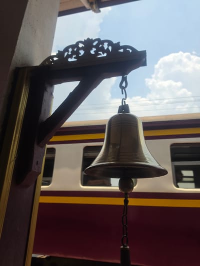 A golden bell hangs from a chain attached to an ornate wooden bracket. The bell is positioned against a backdrop of a train car with a burgundy stripe. The image was taken in Phra Nakhon Si Ayutthaya City Municipality, Thailand.