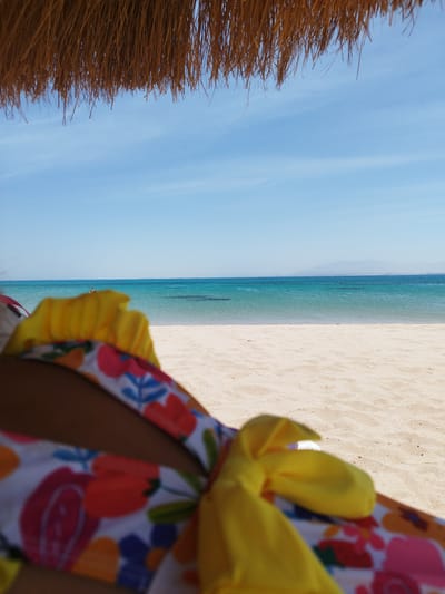 A person is lying on a beach chair on a sandy beach at Ras Abu Soma, Egypt. The person is wearing a floral swimsuit with a yellow tie. The beach is bordered by turquoise water and the horizon is visible in the distance. A thatched roof provides shade in the upper part of the frame.