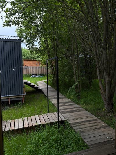 The image displays a serene outdoor scene, likely a private garden or yard, rich with natural elements and functional structures. In the foreground, partially visible on the left, is a dark grey structure constructed from corrugated metal panels, possibly a shed or an outdoor utility building. String lights with spherical bulbs are suspended across the space, originating near this building.

A lush, green lawn dominates the ground level, characterized by thick grass and interspersed with small yellow wildflowers. A winding wooden walkway, composed of horizontal planks, extends from the mid-ground towards the upper right, leading to a set of wooden steps that descend into the grassy area.

The upper section of the image is filled by a dense canopy of trees. Their gnarled, textured branches, adorned with numerous green leaves, create a natural overhead cover. More spherical string lights are visible hanging amongst these branches. In the background, a wooden fence, constructed from horizontal planks, serves as a boundary, with additional trees and greenery beyond it.

The scene is illuminated by soft, diffused daylight, suggesting an overcast sky or filtered sunlight. No people are present, lending a quiet and peaceful atmosphere to the environment. There is no visible text in the image.