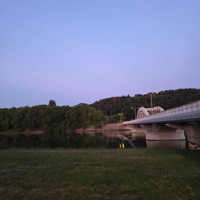 The image depicts a wide shot of the Kolonádový bridge in Piešťany, Slovakia, at dusk. The sky is a soft gradient of blue and purple, transitioning to a darker hue at the top. Below the sky, a dense line of trees runs along the bank of a calm body of water, likely the Váh River. The trees are dark green, reflecting softly in the water, which also shows faint reflections of lights from the bridge and the surrounding area.

The Kolonádový bridge dominates the right half of the image, its concrete arches and walkway spanning across the water. The bridge is illuminated by streetlights, casting a warm glow. The walkway features a metal railing. Some graffiti is visible on the bridge's support structure on the right side. In the foreground, a grassy embankment slopes down towards the water, covered in dark green grass with some patches of lighter, dry grass. The overall scene is serene and tranquil, capturing the quiet beauty of the city at twilight. No people are visible in the image.