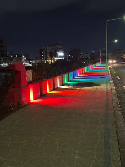 This nighttime image depicts a vibrant walkway illuminated by a spectrum of colorful LED lights. The lights are embedded along the edge of the walkway, creating a rainbow effect that casts a colorful glow on the textured asphalt surface and a low concrete barrier. The walkway appears to be next to a road, indicated by the presence of a street light and what looks like a yellow road sign in the upper portion of the image.

In the background, buildings with illuminated windows suggest an urban or developed area. The sky is dark, indicating it's nighttime. The overall mood is festive and modern, with the colorful lighting creating an inviting and visually striking atmosphere. There are no people visible in the image.

On a red structure in the foreground, there appears to be some text in Korean, though it is largely illegible due to the lighting and angle. The context of Jeju, South Korea, suggests this might be a scenic or recreational area. The weather appears to be clear, as the lights are sharp and there is no visible precipitation.