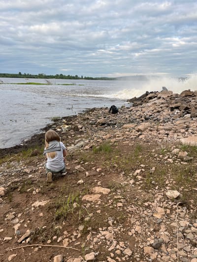 A young child with blonde hair, facing away from the camera, kneels on a rocky, muddy shoreline next to a large body of water. The child is wearing a light-colored t-shirt and dark pants. In the background, waves crash against a rocky embankment. The water appears choppy, and the sky is overcast with clouds. A backpack is visible near the embankment. The scene is located on the shore of the Ufa River in the city of Chernushka, Russia.