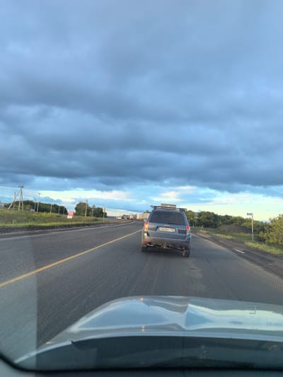 The image shows a view from inside a vehicle driving on a highway under a cloudy sky. A dark blue SUV is visible in front, traveling in the same direction. The highway has a yellow dividing line. The surrounding environment includes grassy areas, trees, and power lines on the left side of the road. In the distance, several trucks are lined up, suggesting a traffic jam or a rest stop. A sign with red lettering is partially visible on the left side of the road. The sky is filled with heavy, dark clouds with some patches of lighter sky visible towards the horizon. The location appears to be a rural or semi-rural highway in Russia, possibly near the city of Valy, as suggested by the prompt.
