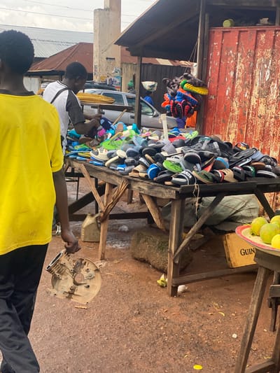In Barakin, Nigeria, a street vendor displays a large assortment of colorful sandals and flip-flops for sale. The shoes are neatly stacked on wooden shelves, forming a vibrant display against a weathered red corrugated metal wall. Several pairs of sandals, including some with athletic-style soles and brand names like "Adidas," are prominently featured.
Two men are visible in the foreground. One, wearing a yellow shirt, appears to be a customer or passerby. The other, in a grey and white shirt, is engaged with a mechanical object. This object, a dirty, beige-colored engine part, is being held by the man, suggesting a repair or exchange.
Behind the shoe stall, a silver vehicle is parked, and the ground is covered with reddish-brown dirt, typical of the local environment. A crate with "GUIN" partially visible on its side and some mangoes on a plate add to the market atmosphere. The scene appears to be taking place outdoors during the day, with natural light illuminating the area. The overall impression is that of a bustling local market where everyday goods are readily available.