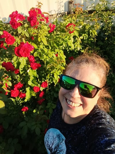 A medium shot selfie shows a smiling woman with blonde hair, wearing black sunglasses and a dark t-shirt with a blue and white pattern of a bare tree against a moon. She is standing in front of a rose bush with bright red flowers and green leaves, with a white wooden fence in the background. The lighting suggests it is daytime, possibly late afternoon, with sunlight casting shadows. The image appears to have been taken outdoors, likely in a garden or yard, and the location is identified as Verea, Russia. The reflection in the sunglasses shows the woman holding a mobile phone, suggesting she is taking the photo herself.