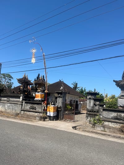 The image captures a scene on a sunny day in Jembrana, Indonesia, likely along a roadside in a residential or temple area. In the foreground, a grey asphalt road is visible, with a concrete curb and sidewalk. Behind the sidewalk is a stone wall, intricately carved and decorated with what appear to be traditional Balinese architectural elements. Two ornate, multi-tiered temple structures with dark, layered roofs stand behind the wall. These structures are adorned with yellow and white cloths, and tall, slender poles with decorative elements are erected nearby. A large tree with lush green foliage is on the left side of the frame, and in the background, more trees, including palm trees and banana trees, are visible, suggesting a tropical environment. Several overhead power lines crisscross the bright blue sky. There are no people or animals visible in the frame, and the overall impression is one of a quiet, culturally significant location.
