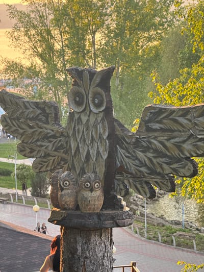 A detailed, carved, wooden sculpture of an owl is the central element of the image. The owl has widespread wings and two smaller, carved owls are positioned on its right side. The sculpture is mounted on a tree trunk base. In the background, blurred scenery depicts trees, a walkway, and some buildings. Indications suggest this is a public area, possibly a park or garden, in Krasny Klyuch, Russia. A person can be seen near the sculpture's base. The overall lighting suggests it is either early morning or late afternoon.