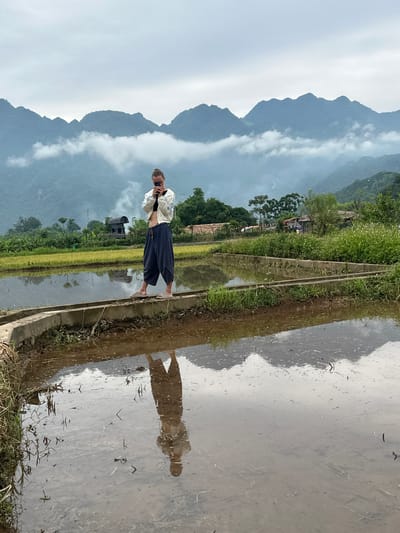 A person stands on a concrete embankment between flooded rice paddies in Mai Chau, Vietnam, holding a mobile phone to take a photo. The person is wearing a white jacket over a black sports bra and loose, dark blue pants. The background features lush green rice fields, trees, and a range of misty mountains under an overcast sky. The water in the paddies reflects the sky, mountains, and the person.