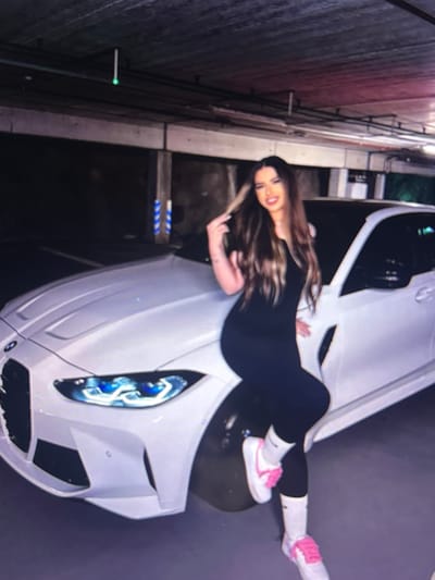 A young woman with long, dark hair and fair skin is posing next to a white BMW car in a dimly lit parking garage. She is wearing a black sleeveless jumpsuit and white socks with pink sneakers. Her right hand is resting on her hip, and her left hand is touching her hair. The car's headlights are illuminated, casting a blue glow. The parking garage appears to be underground, with concrete walls and ceiling. The overall impression is one of a stylish, modern setting. The image is not taken in Beșalma, Moldova.